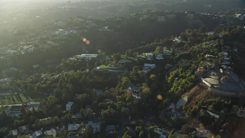 A reverse aerial view of luxurious hillside mansions in Beverly Glen Los Angeles surrounded by hazy forest areas during daytime