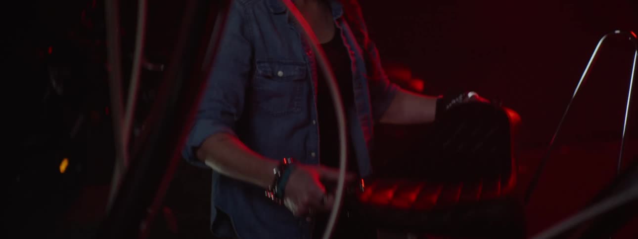 A woman walking over to a motorcycle in a red lit room and takes the seat off and looks at it