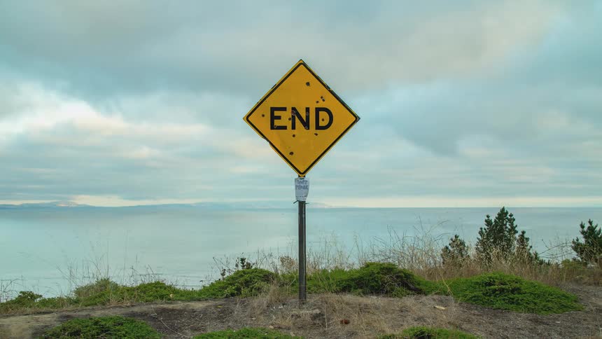 A yellow road sign at the end of a road with the ocean in the background