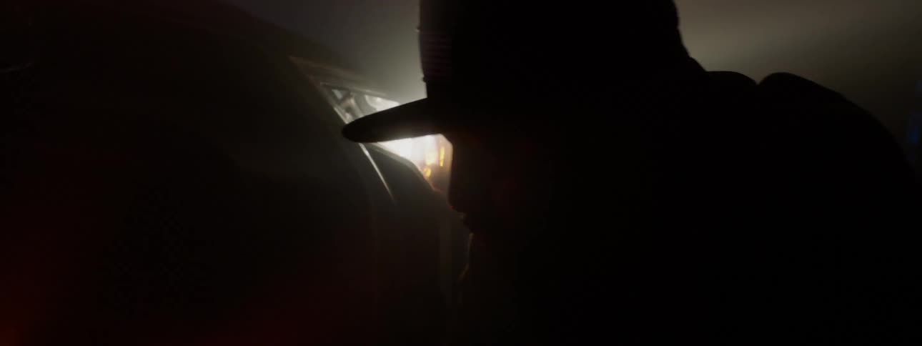 A silhouette of a man wearing a baseball cap kneeling next to a vehicles head lights at night
