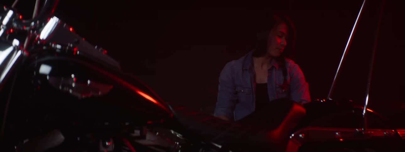 A woman patting a motorcycle seat as she smiles and rolls away on a stool as the engine is shown