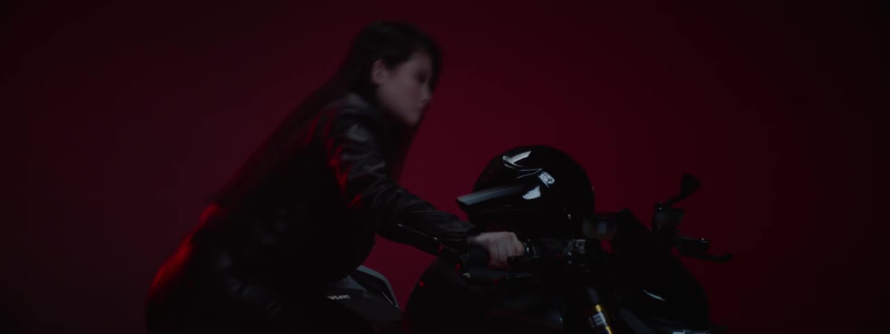 A woman sets her helmet on the motorcycle handlebars as she walks around it and sits on the seat