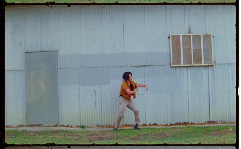 A woman in an orange shirt dancing around in front of a blue building and moving left to right