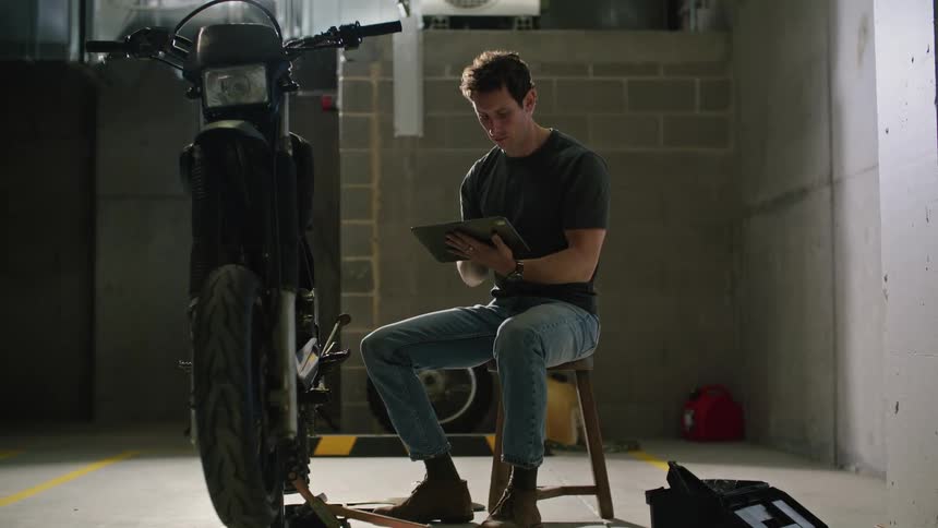 Man sitting on stool uses tablet while examining motorcycle