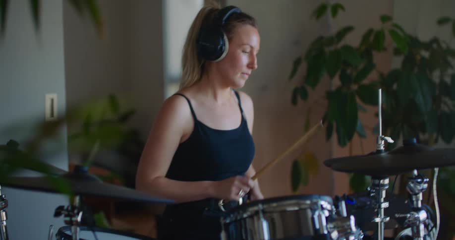 A woman sitting at a drum set hits the drum heads using a pair of drum sticks