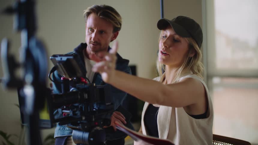 A woman and a man talking as the woman points and shows the man something as she stands in front of a camera