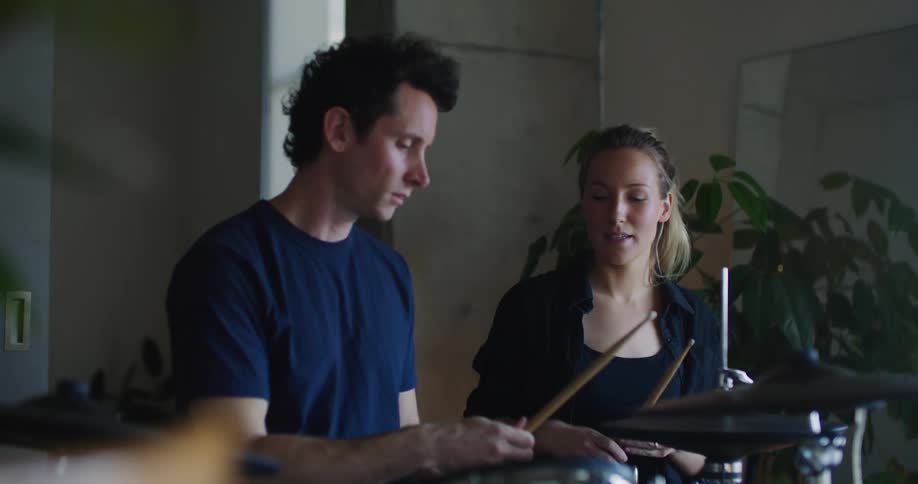 A young blonde woman is teaching a man how to play the drums