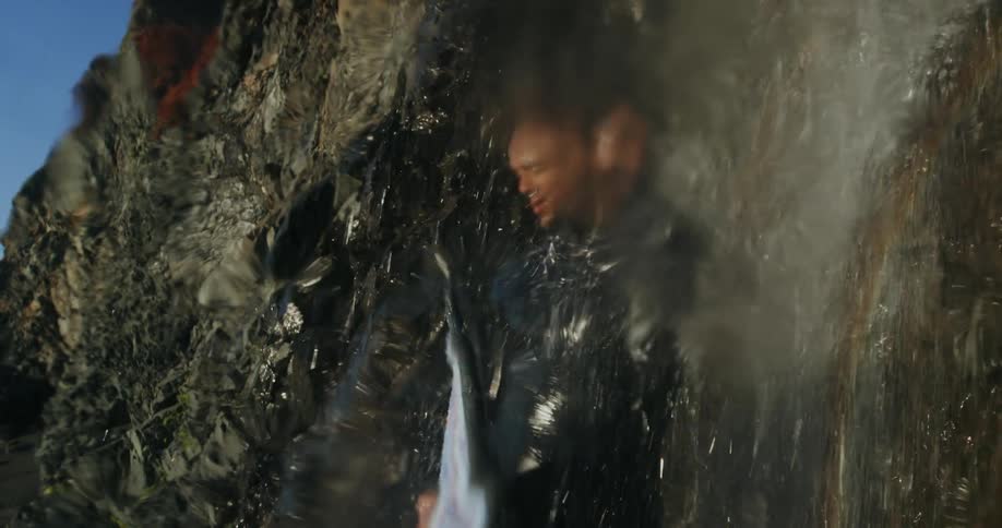 Man with wetsuit and surfboard showers in waterfall