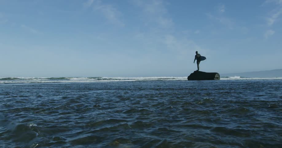 Man with wetsuit stands on rock in sea holding surfboard