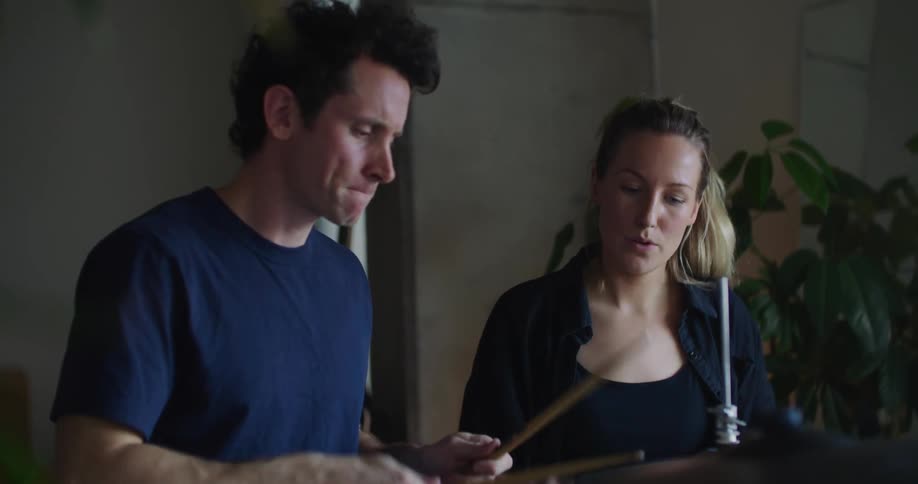 Blonde woman with ponytail and black shirt teaches man wearing dark blue shirt how to play drums