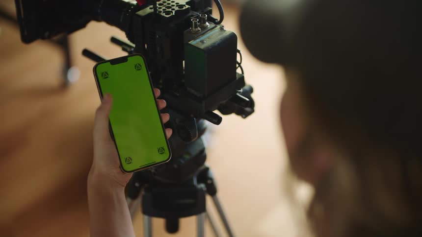 A close up of a woman wearing a baseball cap scrolling on her cell phone next to a camera on a tripod
