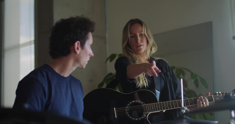A man playing the drums and a woman playing the guitar as the woman stops them and points something out to the man