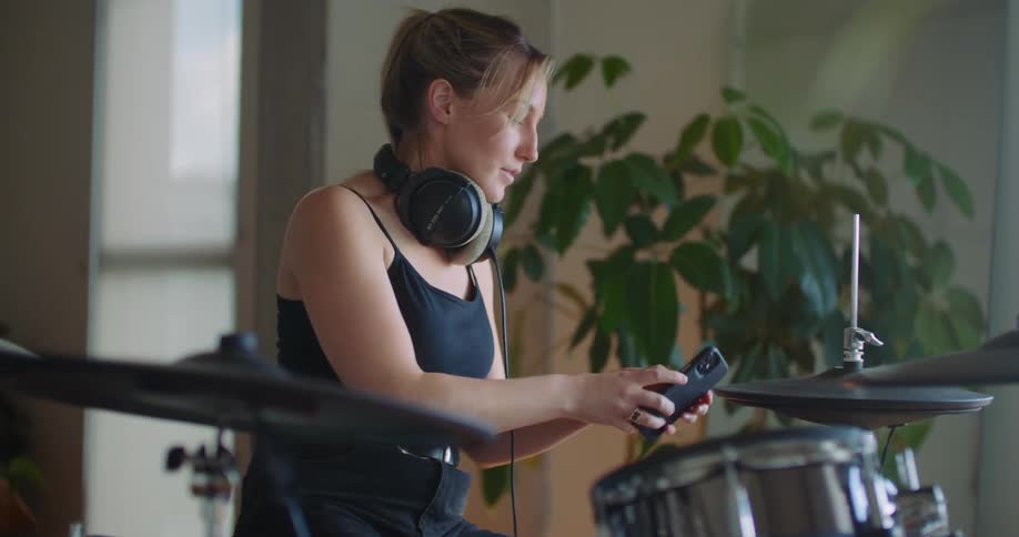 A woman sitting playing a set of drums with headphones on as she stops and answers her cellphone