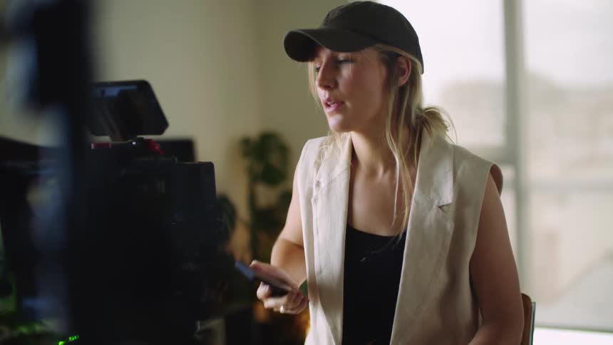 A woman is sitting behind a camera on a tripod as she holds a tablet computer and talks on the phone