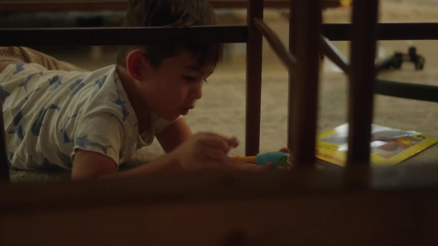 A little boy laying on the carpeted floor playing with toys under a chair