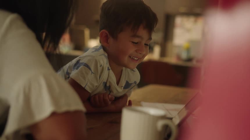 A boy sitting beside a woman at a table smiles while watching the screen of a tablet