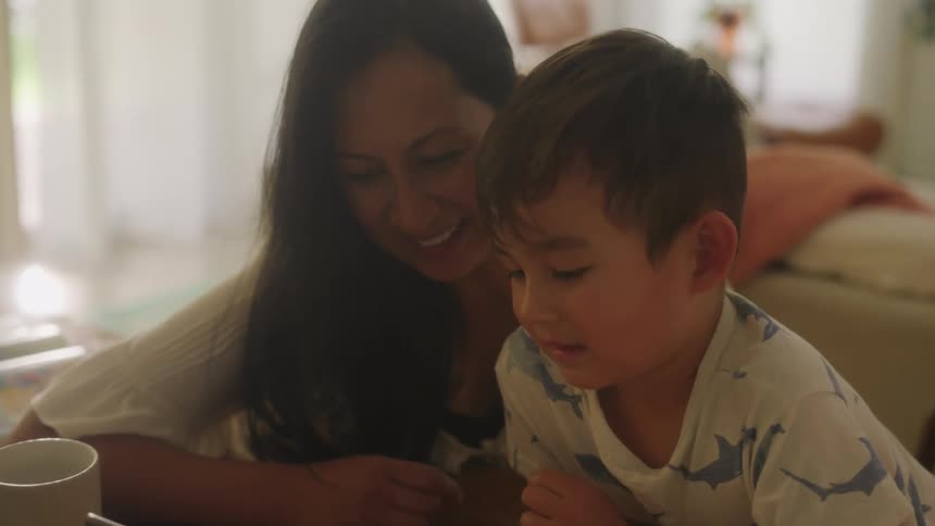 Mother and son sitting together at dining table watching tablet and learning smiling and laughing probably home schooling