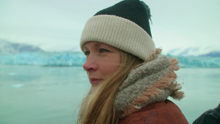 A blonde woman overlooks a cold body of water with a glacier in the background