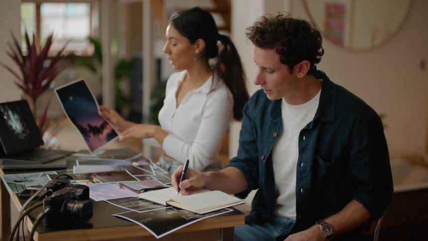 Man in dark blue and white shirt writes down notebook next to woman with ponytail white shirt and jeans holding photographs at desk