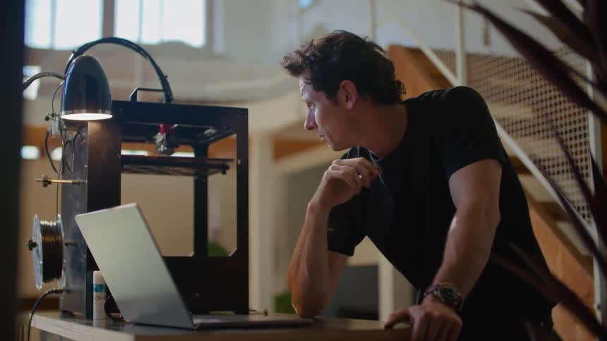 A man using video chat on a laptop checks the 3d printer operating beside him