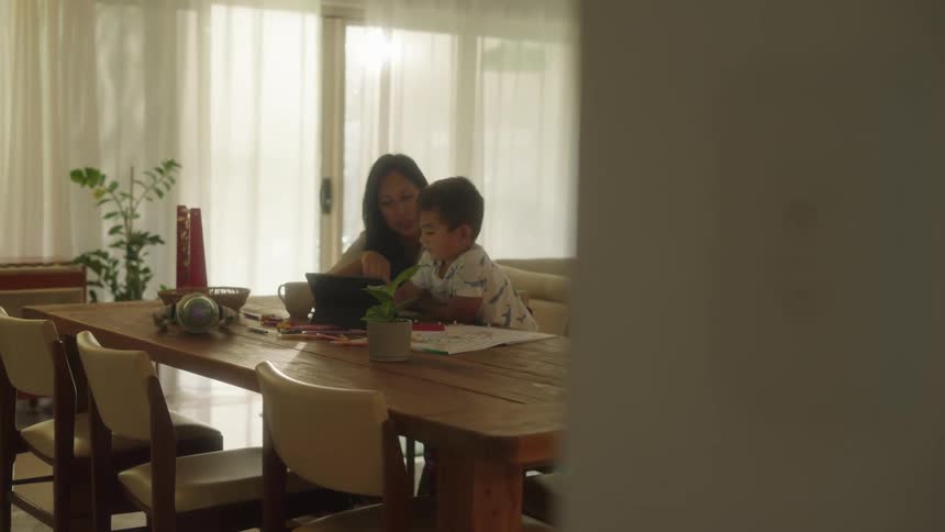 Mother and son sitting together at dining table smiling and laughing looking at tablet and probably homeschooling