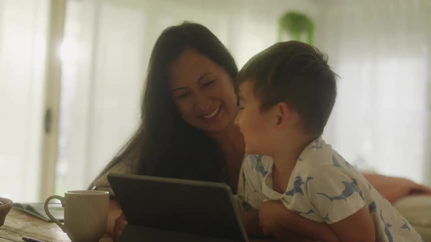 Mother and son watches tablet at table laughing in the living room