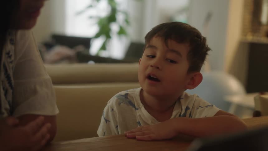 A young boy sitting at a table looking at a tablet next to his mother