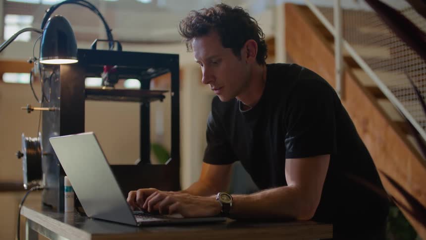 A man is working on a machine and standing over a table as he types on a laptop computer