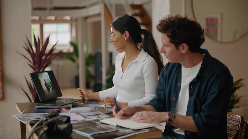 Woman with ponytail and white shirt shows man in dark blue shirt photos on tablet sitting at desk
