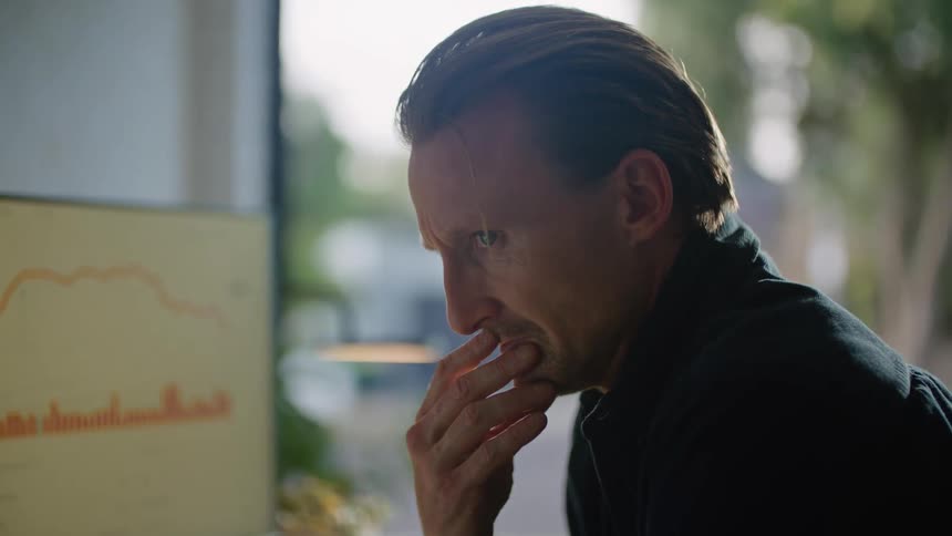 A man is sitting at a desk with a laptop computer and a large monitor with his hand on his face as he thinks
