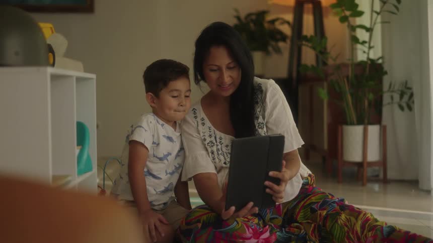 A mother and her son sitting on the floor looking at a tablet smiling and laughing