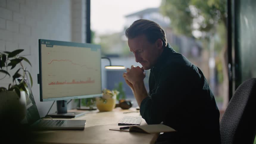 Nervous and sad man in black shirt sits at desk looking at computer and laptop at the office