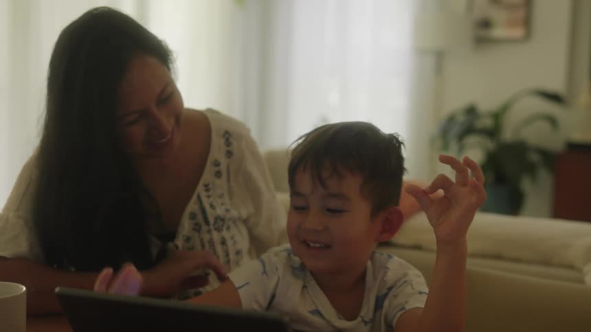 A mother and son sit at a table in front of a tablet as the mother smiles and hugs the boy