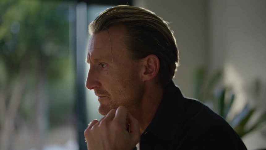 A man is sitting at a desk leaning over a computer keyboard with his hand on his face as he thinks