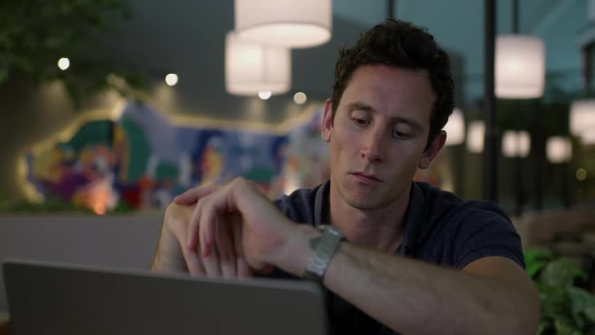 A man in a big fancy office sits in front of a laptop at dusk and quietly checks his watch