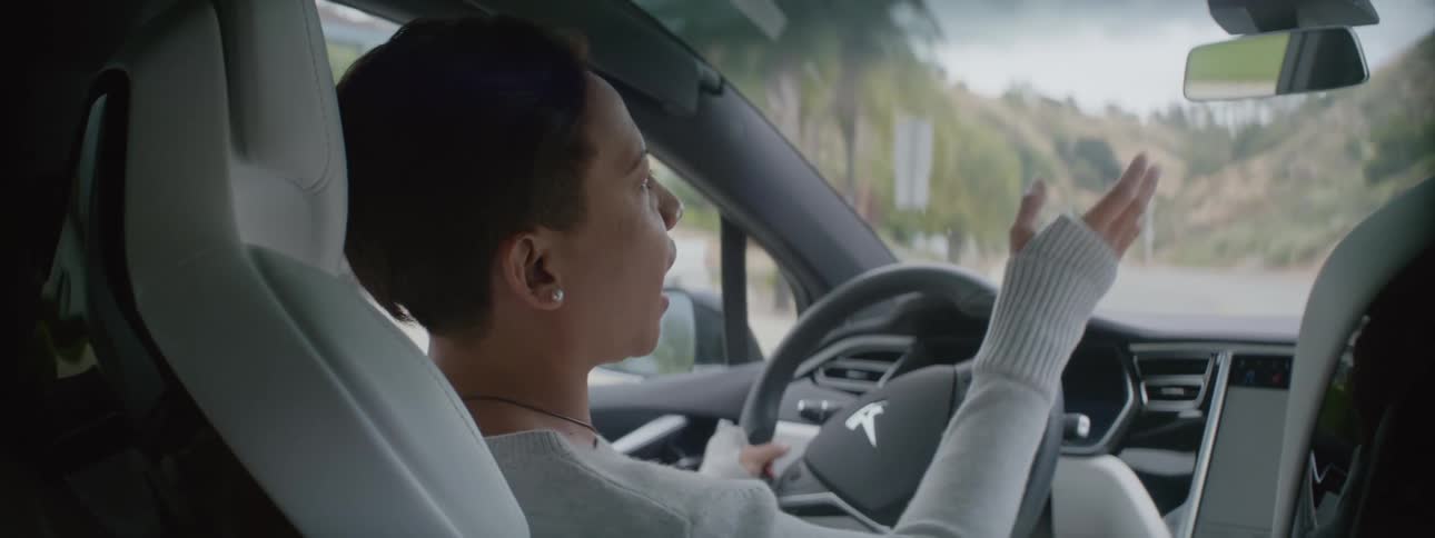 A woman is driving an automobile and she lifts her hands off the steering wheel as she talks