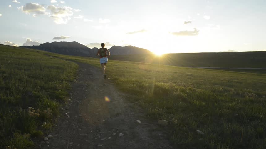 A woman runs down a dirt trail on a grassy plain toward distant mountain peaks