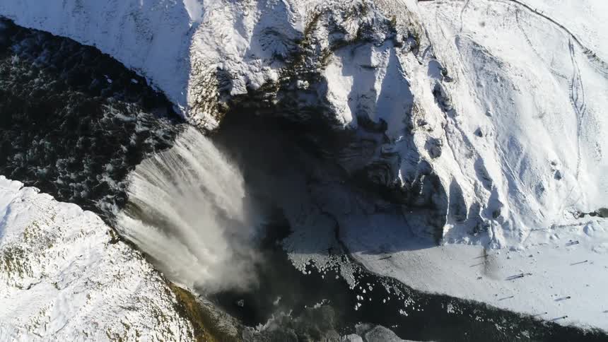 Snowcovered mountains are shown from above with a waterfall flowing into a river between them