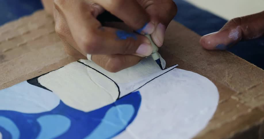 Hands use a black marker to create dark outlines over a painting on a wood board