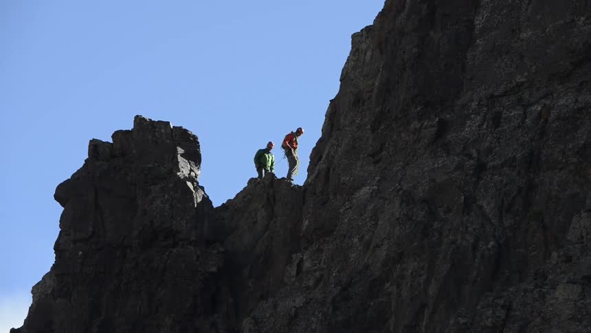 Two men are walking on a rough rocky mountain and one man is carrying a rope