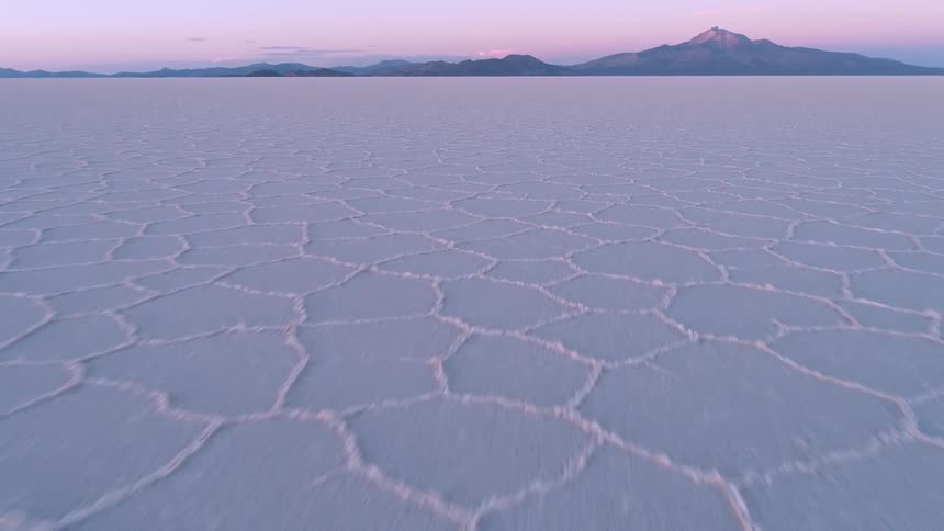 A camera moves across an icy plain toward a distant mountain range at sunset