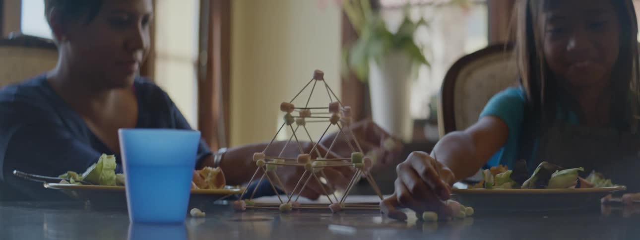 A mother and daughter sit at a table together with plates of food in front of them while they play with sticks and put them together