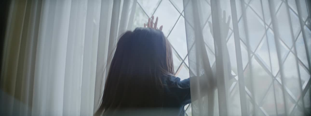 Close up of a girl touching the glass of a window throught the curtains to look out