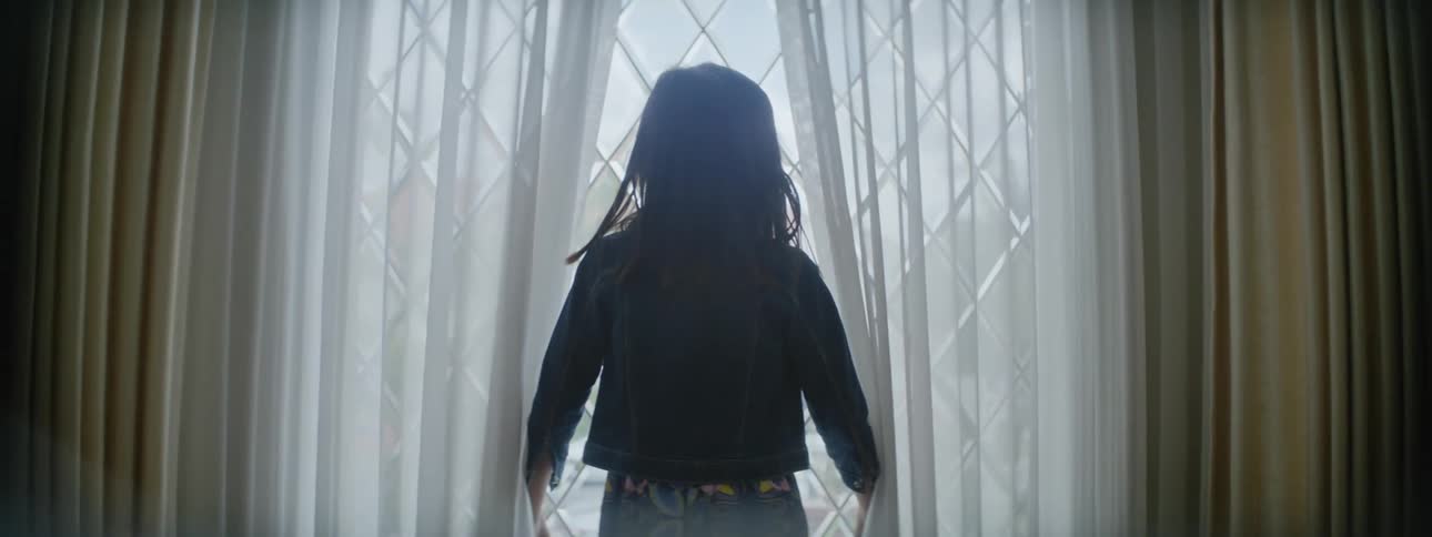 A girl parts the curtains while approaching a window before gazing outside