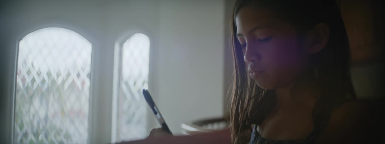 A girl uses a pen and paper to write while sitting inside a house
