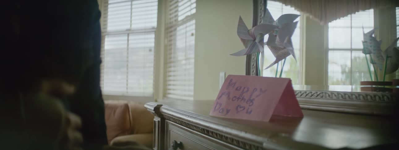 A mother and daughter look at a mothers day card and paper windmills placed on a dresser and hug