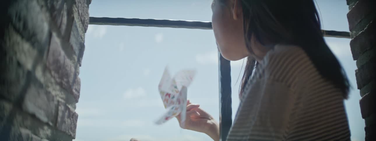 A young girl plays with a paper windmill toy while standing in an open window in the day