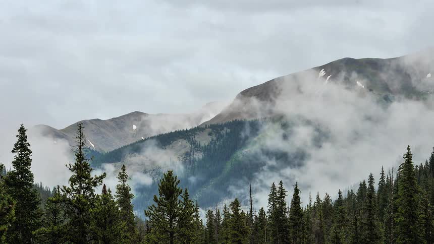 A timelapse view of a cloudy mountain peak above an evergreen forest from day to dusk