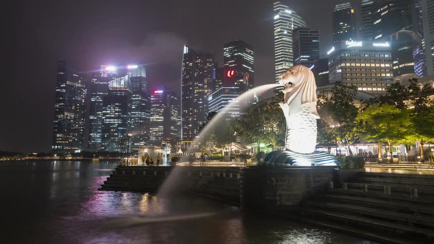 A time lapse of a fountain spraying into a small body of water with tall city buildings lit up at night in the background