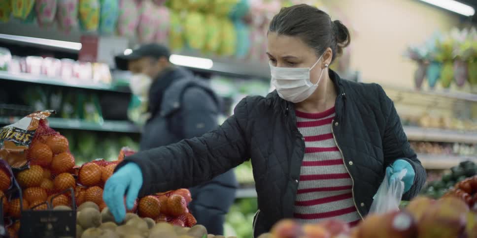 A woman with disposable gloves and a mask putting fruit in a bag